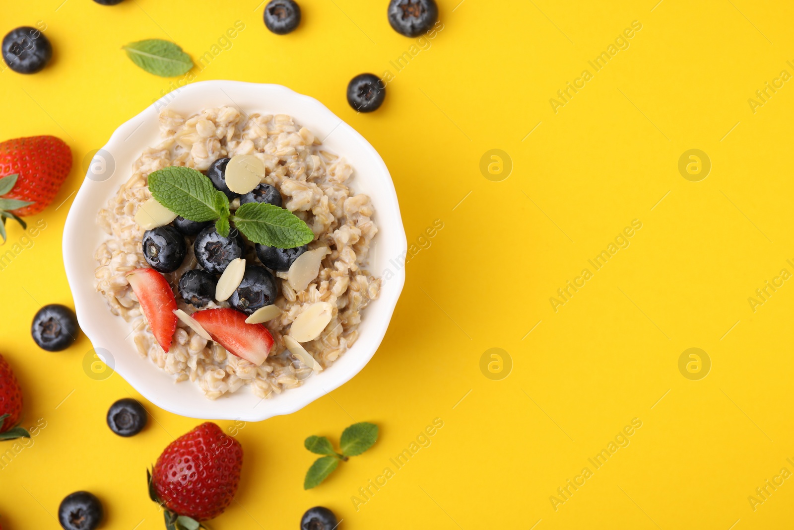 Photo of Tasty oatmeal with strawberries, blueberries and almond petals in bowl surrounded by fresh berries on yellow background, flat lay. Space for text