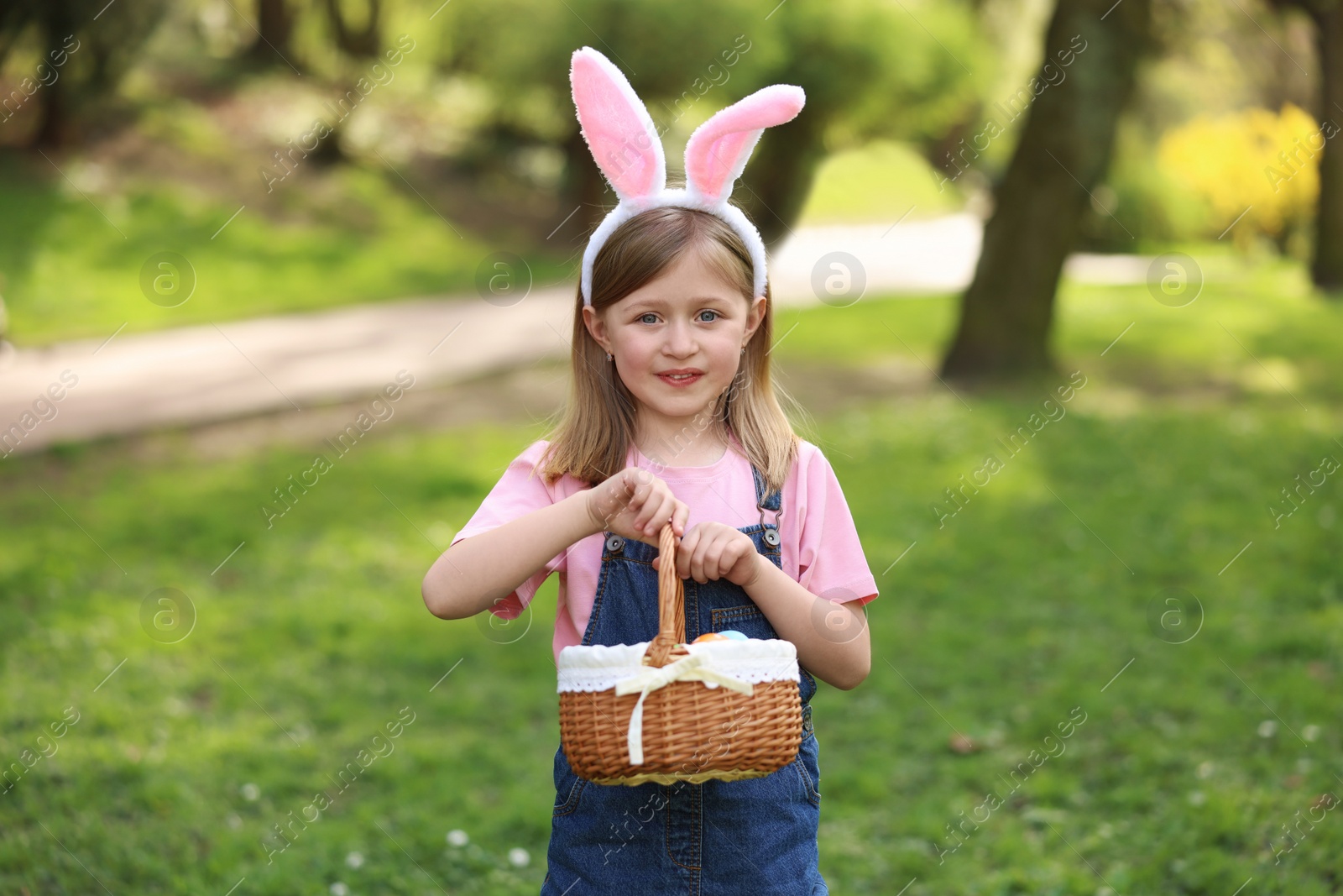 Photo of Easter celebration. Cute little girl with bunny ears holding wicker basket outdoors