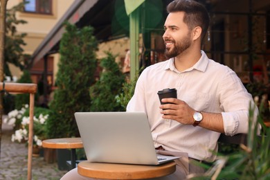 Handsome young man with cup of coffee working on laptop at table in outdoor cafe
