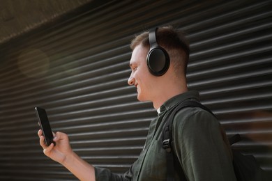 Photo of Smiling man in headphones using smartphone near shutters outdoors