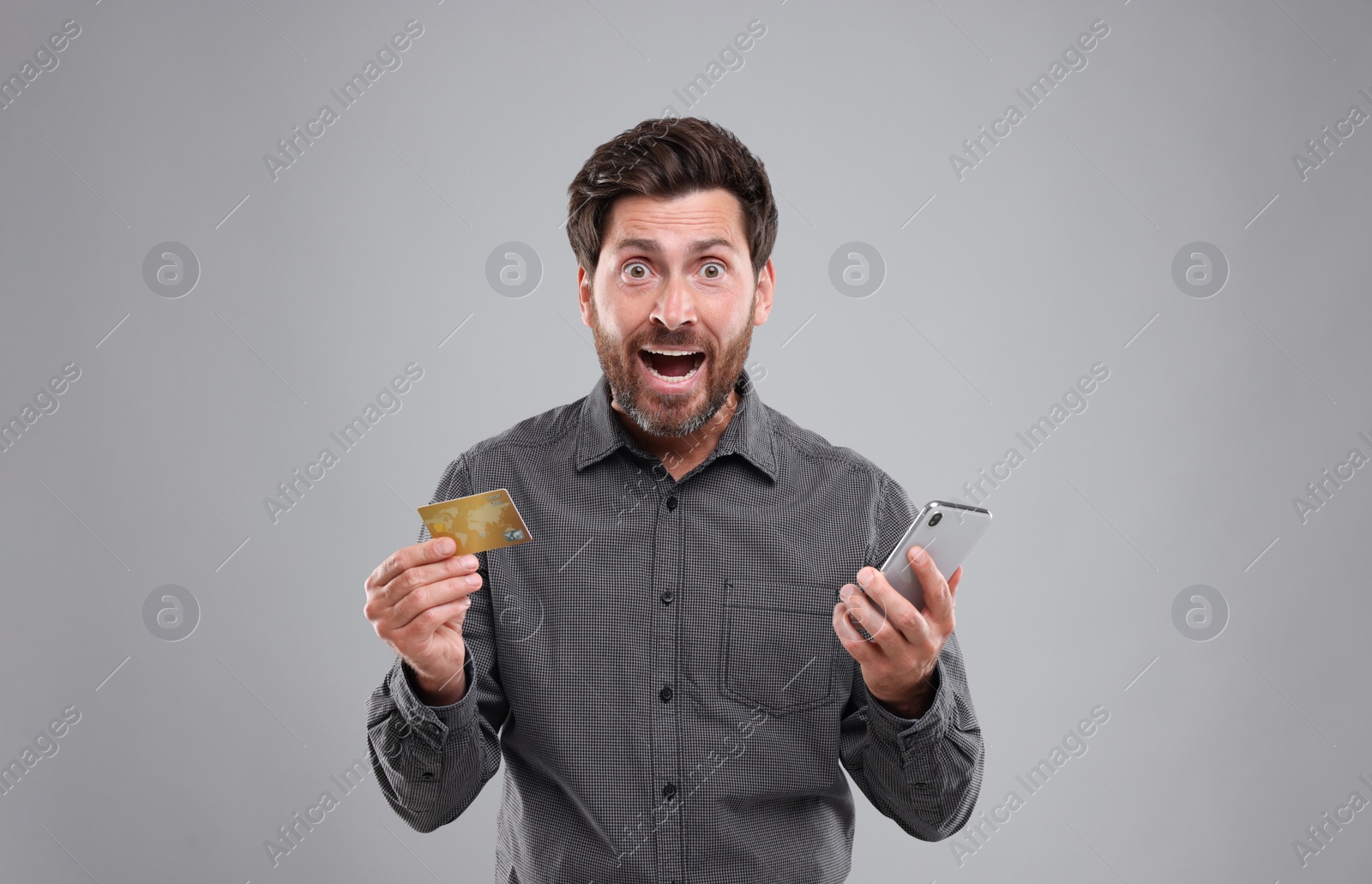 Photo of Emotional man holding smartphone and credit card on light grey background. Be careful - fraud