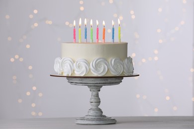 Photo of Birthday cake with burning candles on white table against blurred festive lights