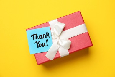 Photo of Gift box and light blue paper note with phrase Thank You on yellow background, top view