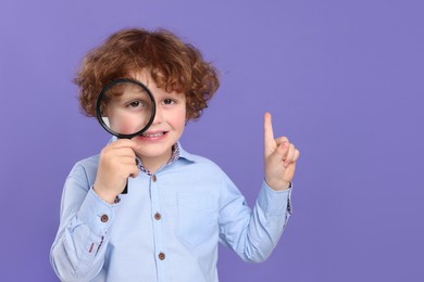 Cute little boy looking through magnifier glass and pointing on violet background, space for text