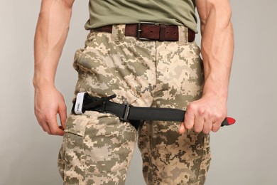 Photo of Soldier in military uniform applying medical tourniquet on leg against light grey background, closeup