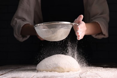 Photo of Woman sprinkling flour over dough at wooden table on dark background, closeup