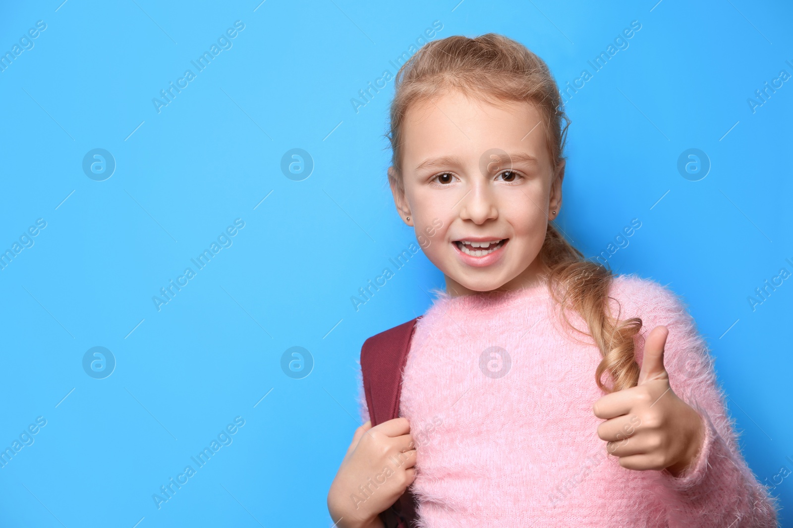 Photo of Happy little girl with backpack showing thumbs up on light blue background. Space for text