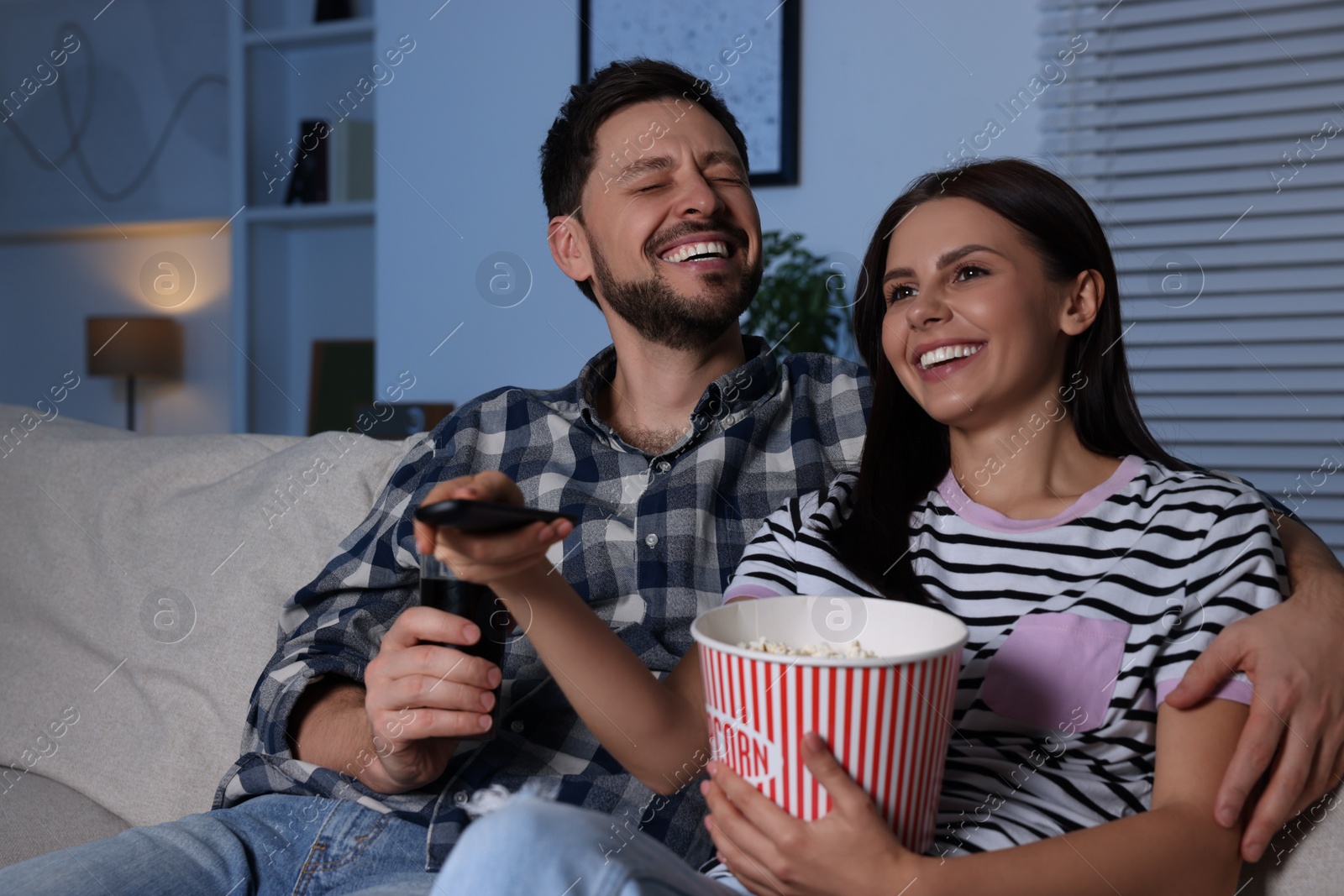 Photo of Happy couple watching show at home in evening. Woman holding popcorn and changing TV channels with remote control
