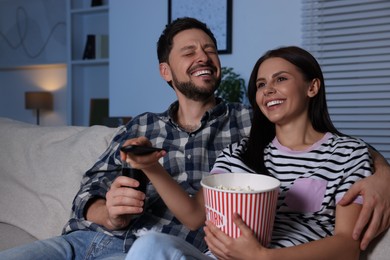 Photo of Happy couple watching show at home in evening. Woman holding popcorn and changing TV channels with remote control