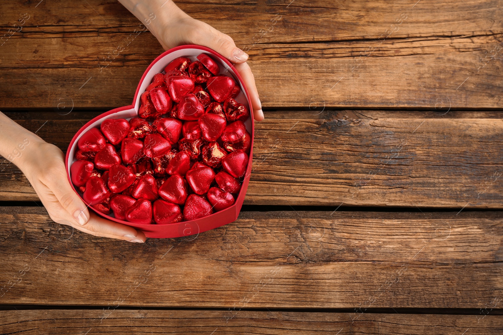 Photo of Woman holding box with heart shaped chocolate candies at wooden table, top view. Space for text