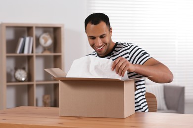 Photo of Happy young man opening parcel at table indoors. Internet shopping