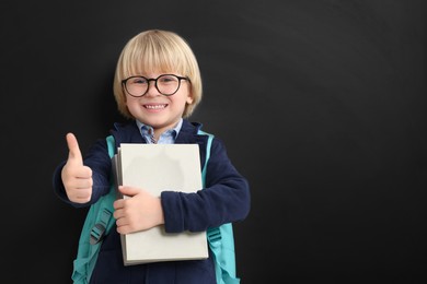 Happy little school child with notebooks showing thumbs up near chalkboard. Space for text