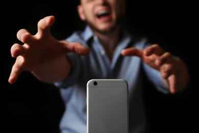 Emotional man reaching for smartphone on black background, closeup. Addiction concept