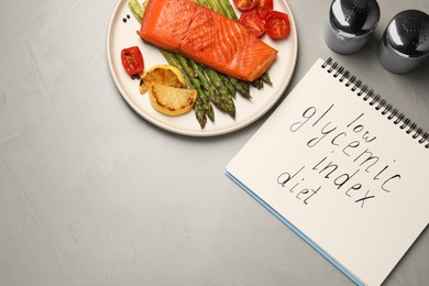 Photo of Notebook with words Low Glycemic Index Diet and plate of tasty grilled salmon on grey table, flat lay. Space for text