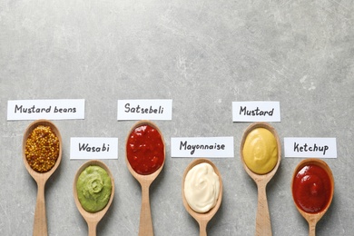 Photo of Different sauces in spoons and name tags on gray background, flat lay