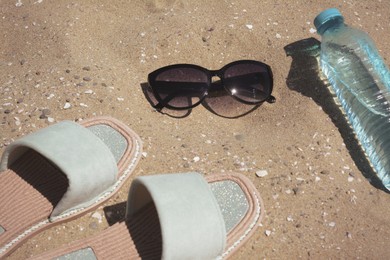 Photo of Stylish sunglasses, slippers and bottle of water on sand