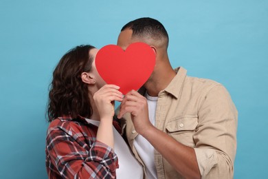 Photo of Lovely couple kissing behind red paper heart on light blue background. Valentine's day celebration