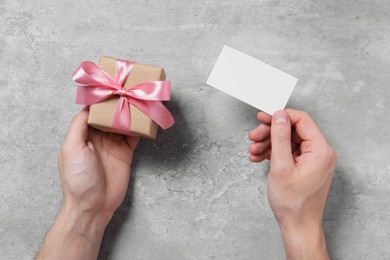 Photo of Man holding gift box with pink bow and blank greeting card at grey table, top view