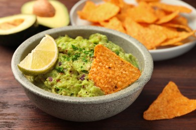 Photo of Bowl of delicious guacamole, lime and nachos chips on wooden table, closeup