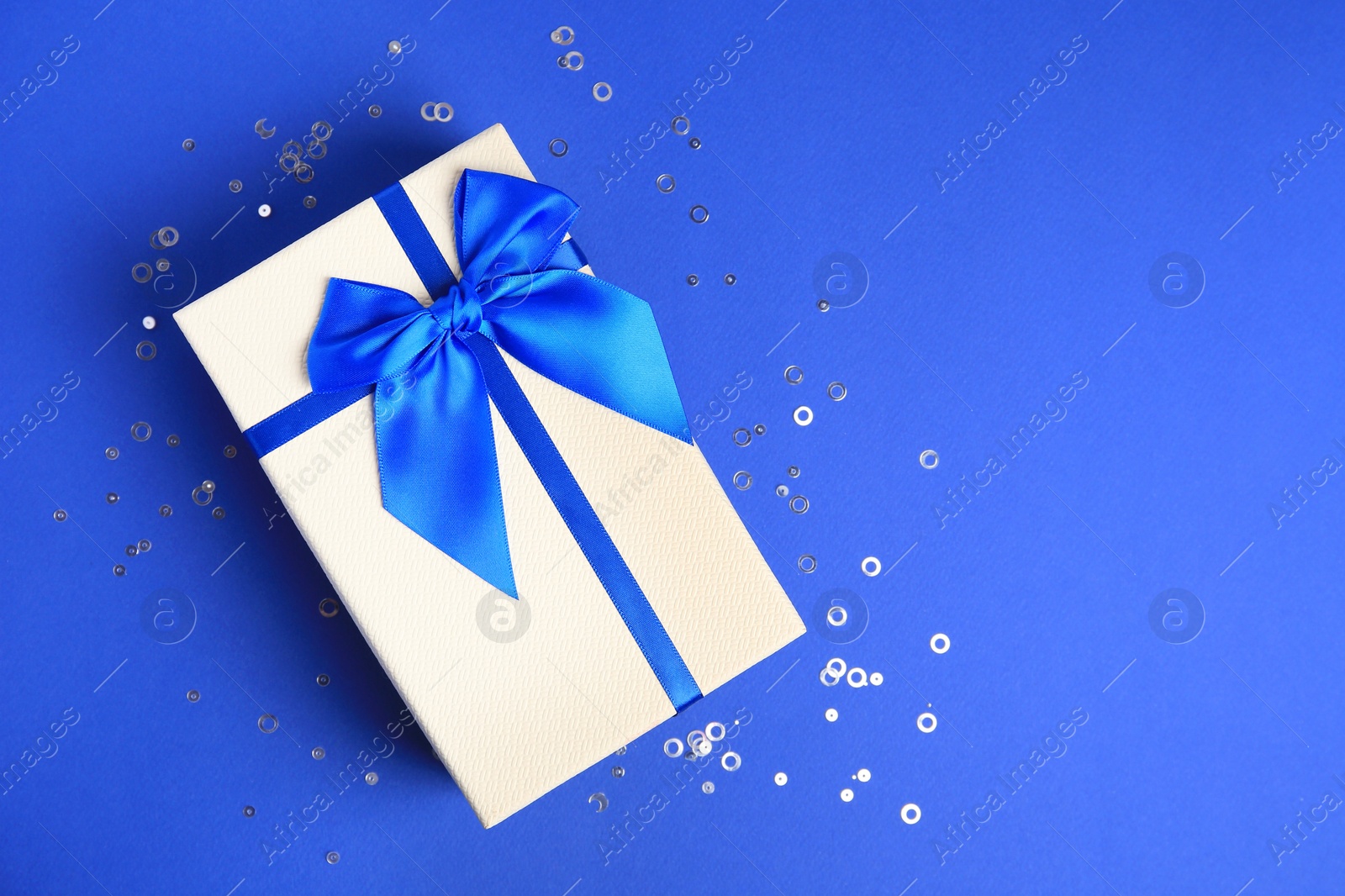 Photo of Beautiful gift box with bow and confetti on blue background, top view. Space for text