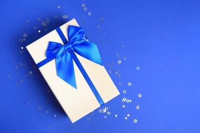 Beautiful gift box with bow and confetti on blue background, top view. Space for text