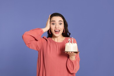 Photo of Coming of age party - 21st birthday. Surprised woman holding delicious cake with number shaped candles on violet background