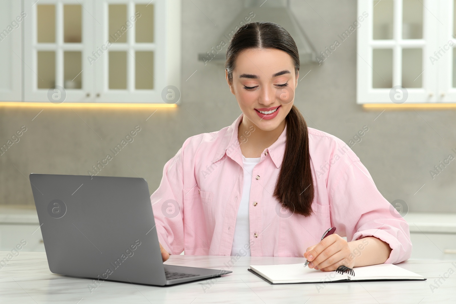 Photo of Woman writing something in notebook while using laptop at white table in kitchen