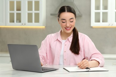 Woman writing something in notebook while using laptop at white table in kitchen