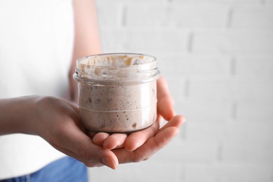Woman holding glass jar with fresh sourdough starter on blurred background, closeup. Space for text