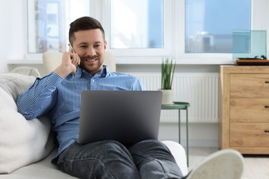 Photo of Happy man talking on phone while working with laptop on sofa at home