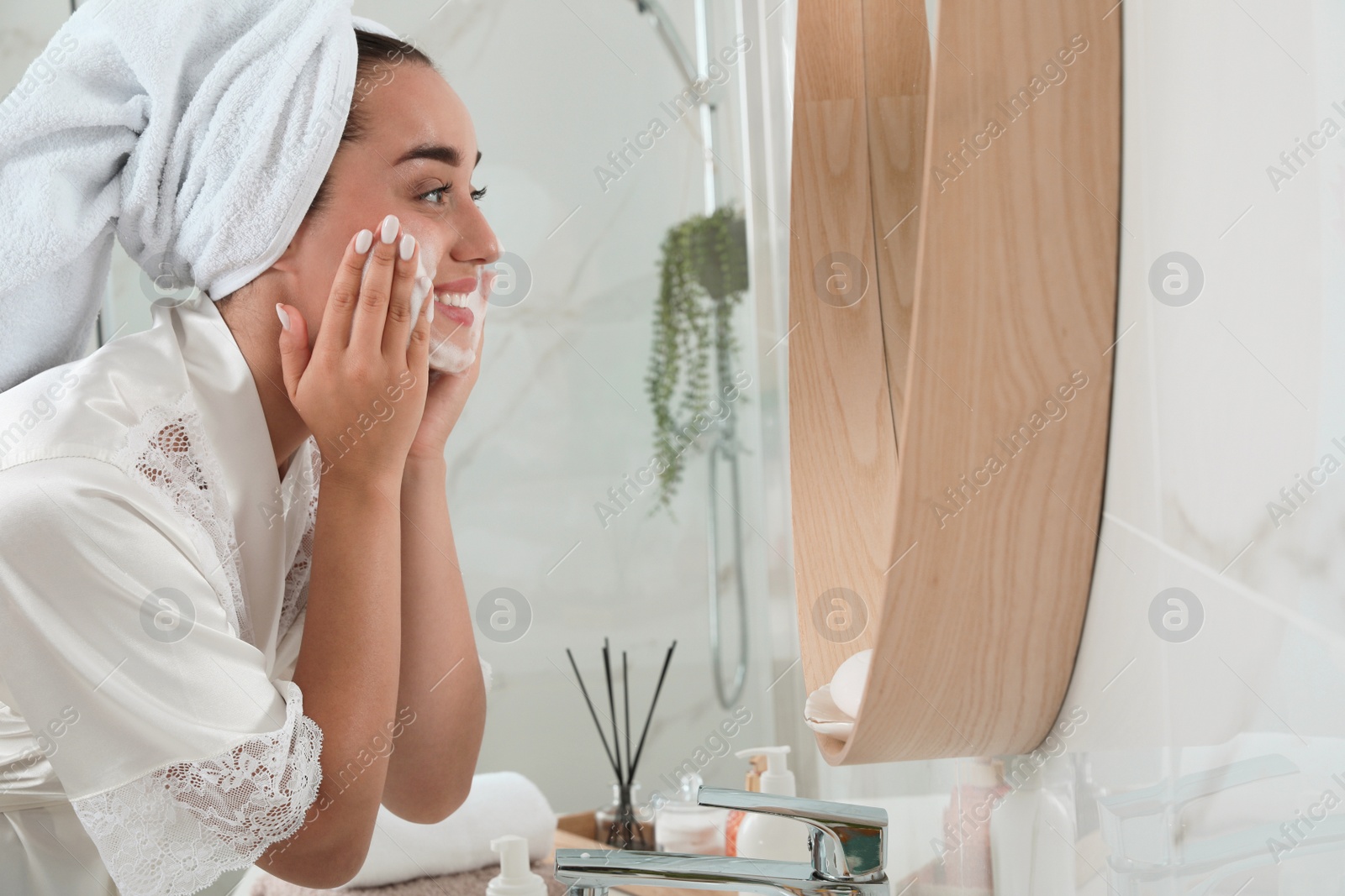 Photo of Beautiful young woman applying cleansing foam onto face near mirror in bathroom, space for text. Skin care cosmetic