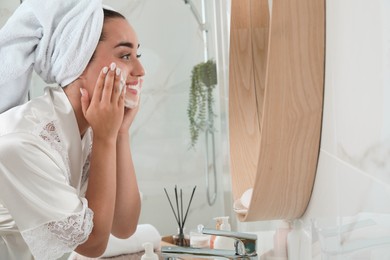 Photo of Beautiful young woman applying cleansing foam onto face near mirror in bathroom, space for text. Skin care cosmetic