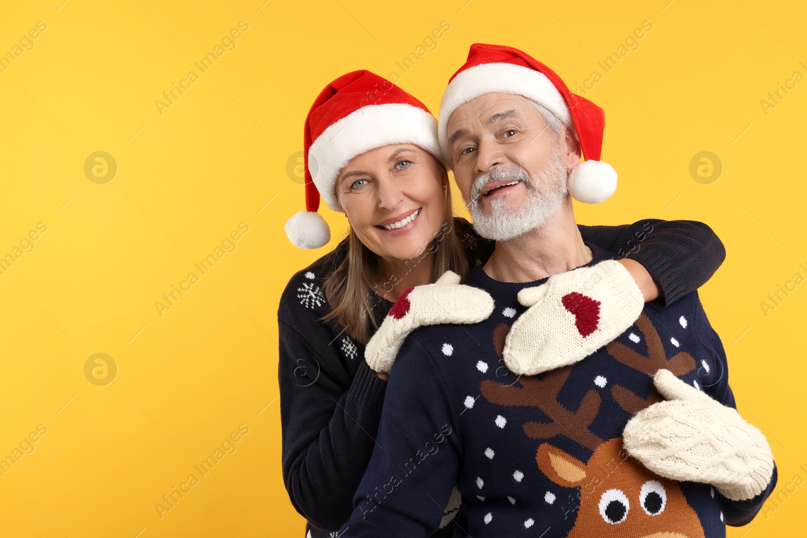 Photo of Happy senior couple in Christmas sweaters, Santa hats and knitted mittens on orange background. Space for text