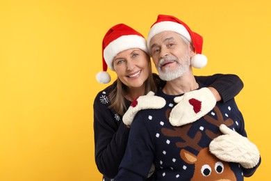 Photo of Happy senior couple in Christmas sweaters, Santa hats and knitted mittens on orange background. Space for text