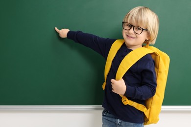 Happy little school child with backpack pointing at chalkboard. Space for text