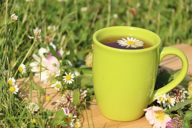 Green cup with tea, different wildflowers and herbs on wooden board in meadow, closeup