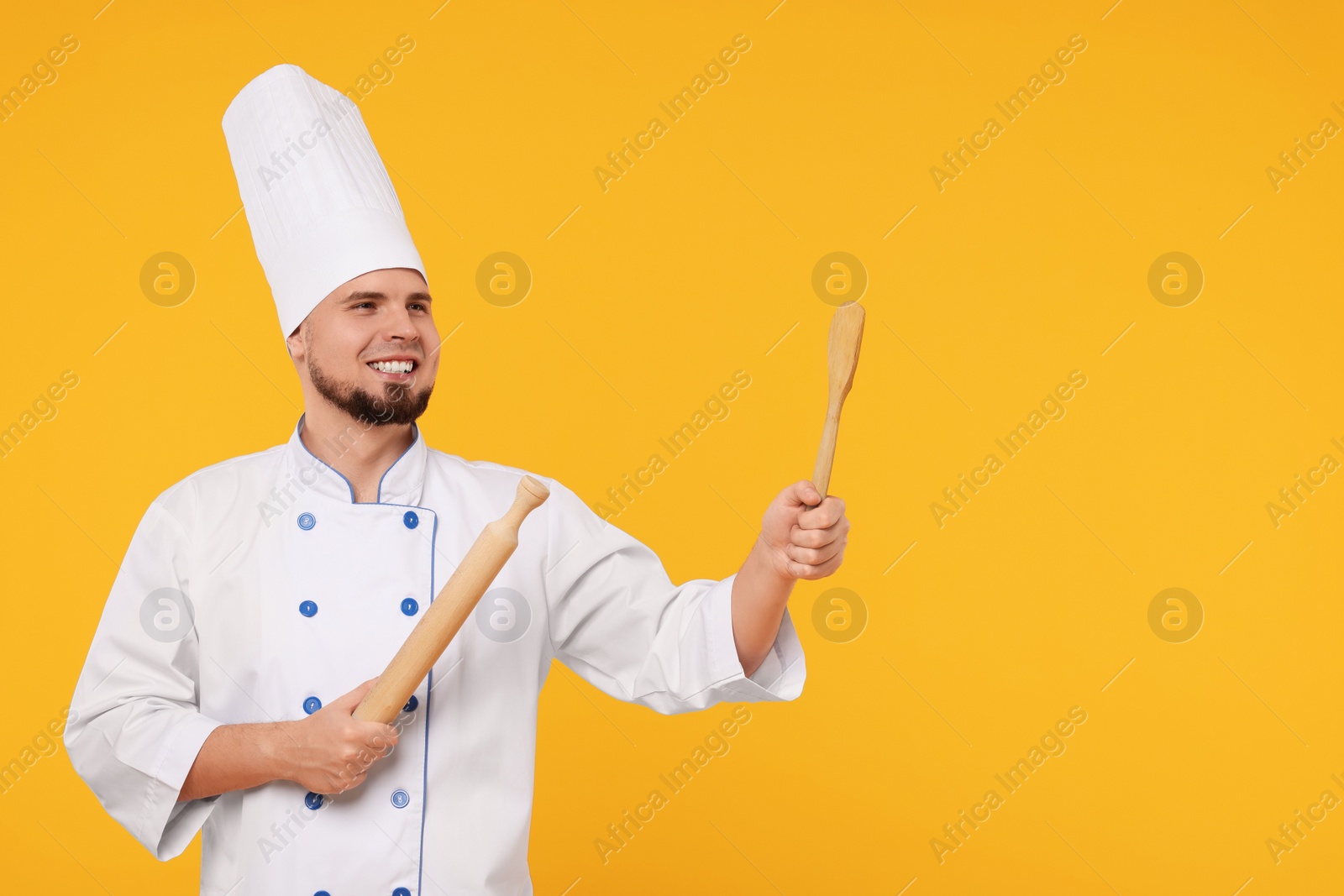 Photo of Happy professional confectioner in uniform holding rolling pin and spatula on yellow background. Space for text