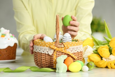 Closeup of woman putting painted egg into Easter basket at white marble table