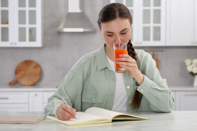 Photo of Woman drinking juice while writing in notebook at white marble table indoors
