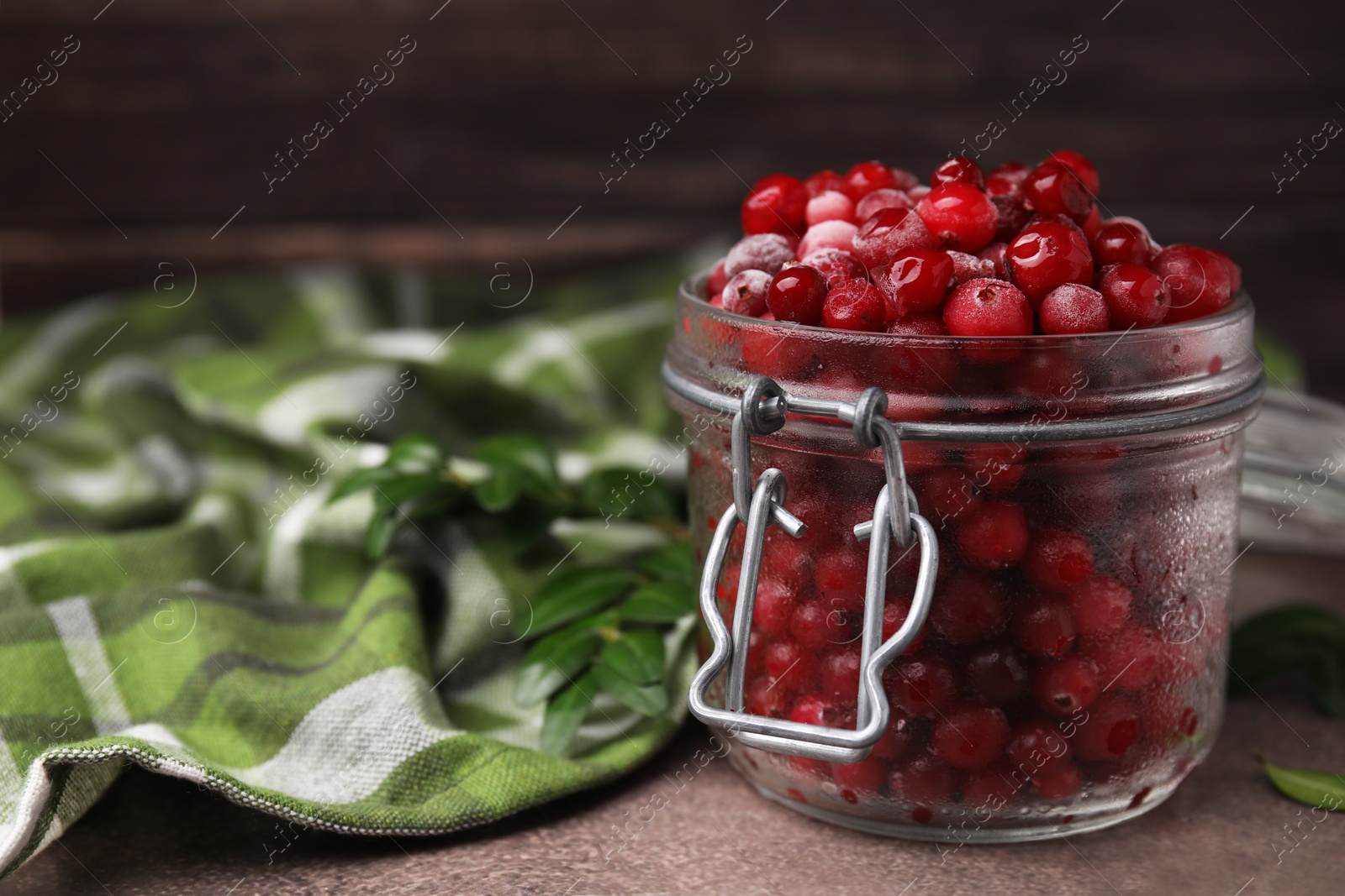 Photo of Frozen red cranberries in glass jar and green leaves on brown textured table, closeup. Space for text