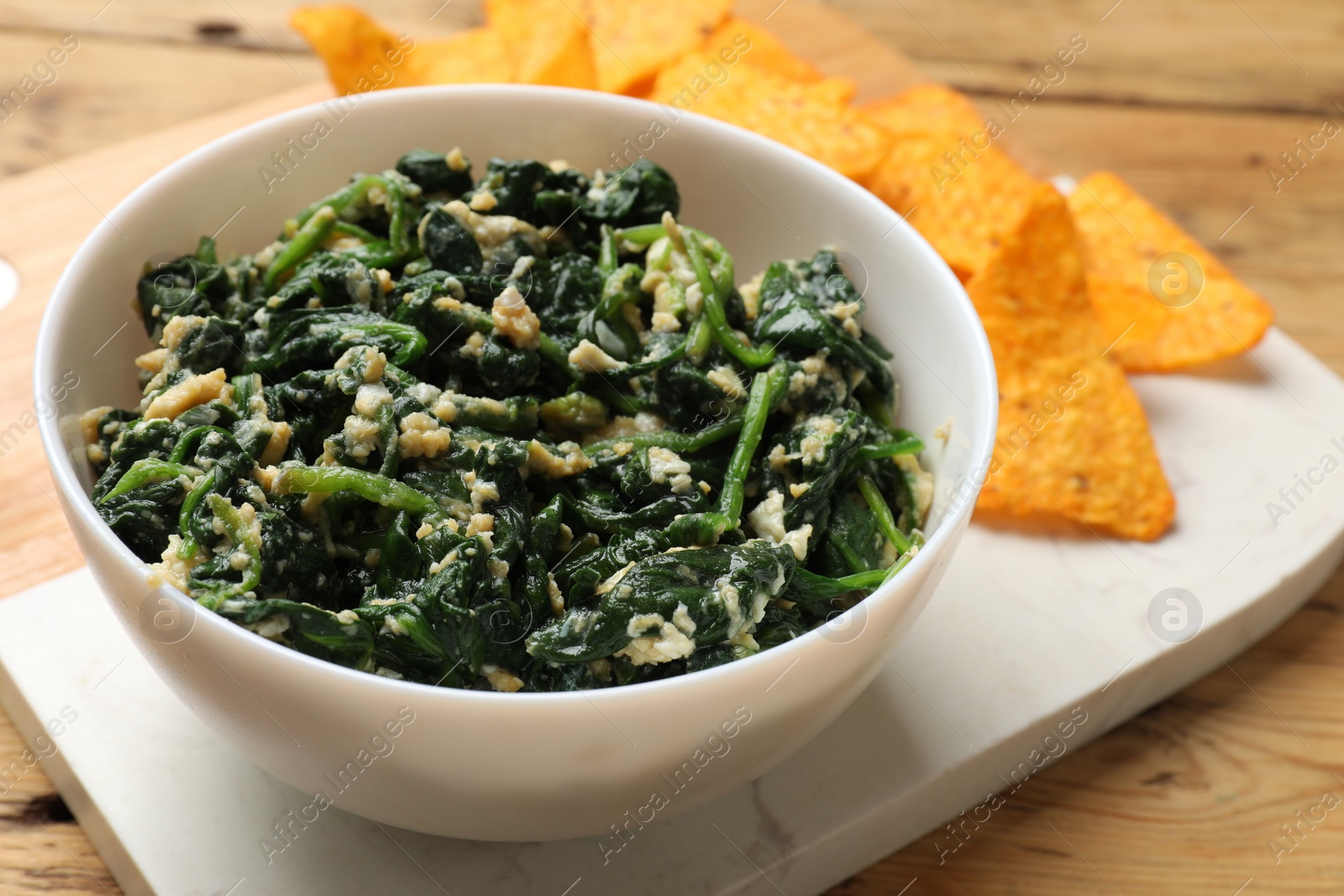 Photo of Tasty spinach dip with eggs in bowl and nachos chips on wooden table, closeup