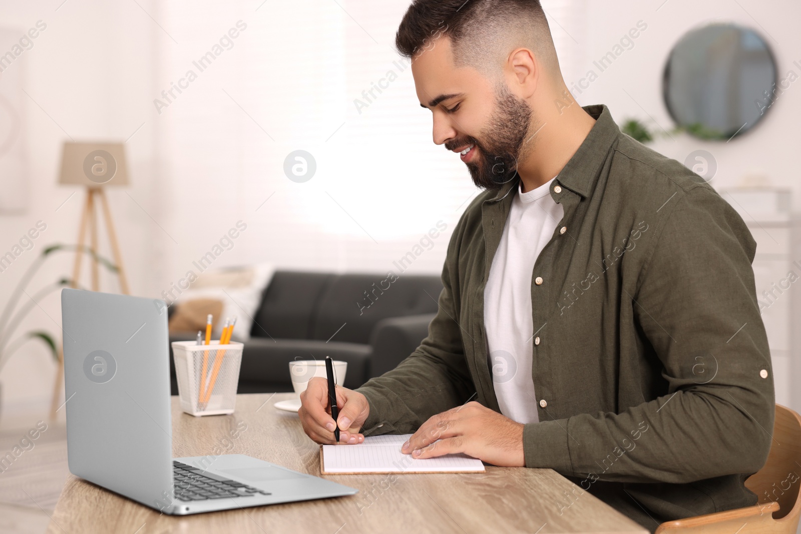 Photo of Young man writing in notebook at wooden table indoors