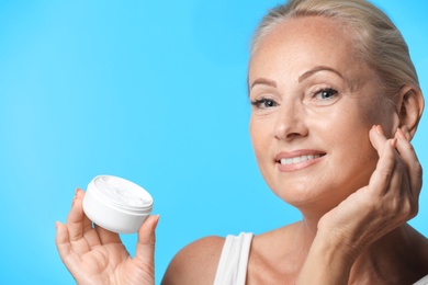 Portrait of beautiful mature woman with perfect skin holding jar of cream on light blue background