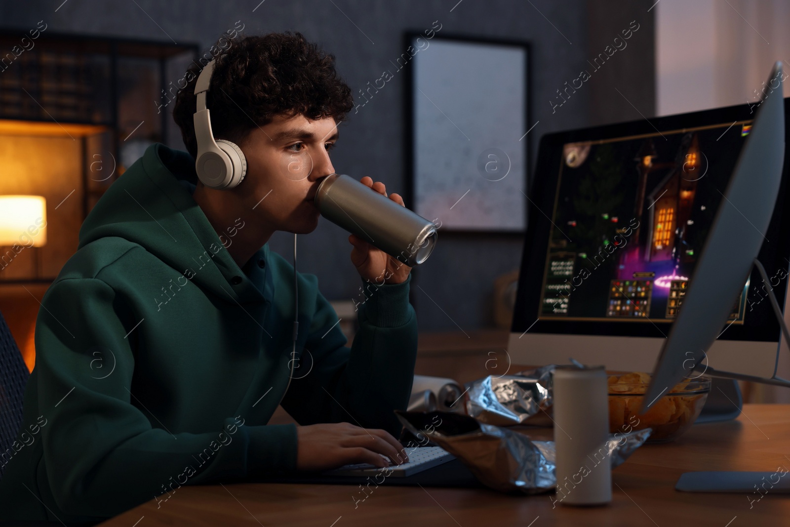 Photo of Young man with energy drink and headphones playing video game at wooden desk indoors