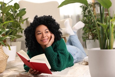 Photo of Relaxing atmosphere. Woman holding book and dreaming surrounded by potted houseplants at home