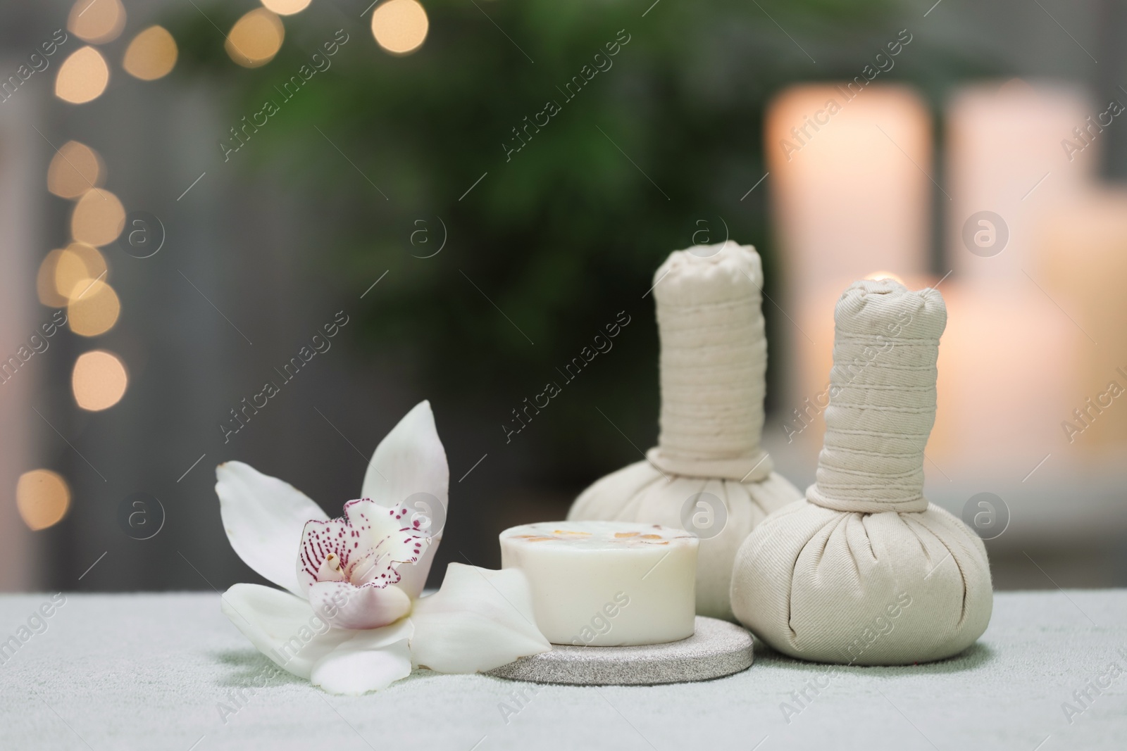 Photo of Spa composition. Herbal massage bags, soap bar and orchid flower on white towel against blurred lights, space for text