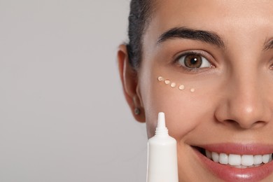 Photo of Beautiful young woman applying gel on skin under eye against light grey background, closeup. Space for text