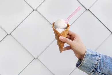 Photo of Woman holding delicious ice cream in waffle cone with plastic spoon near white wall, closeup. Space for text