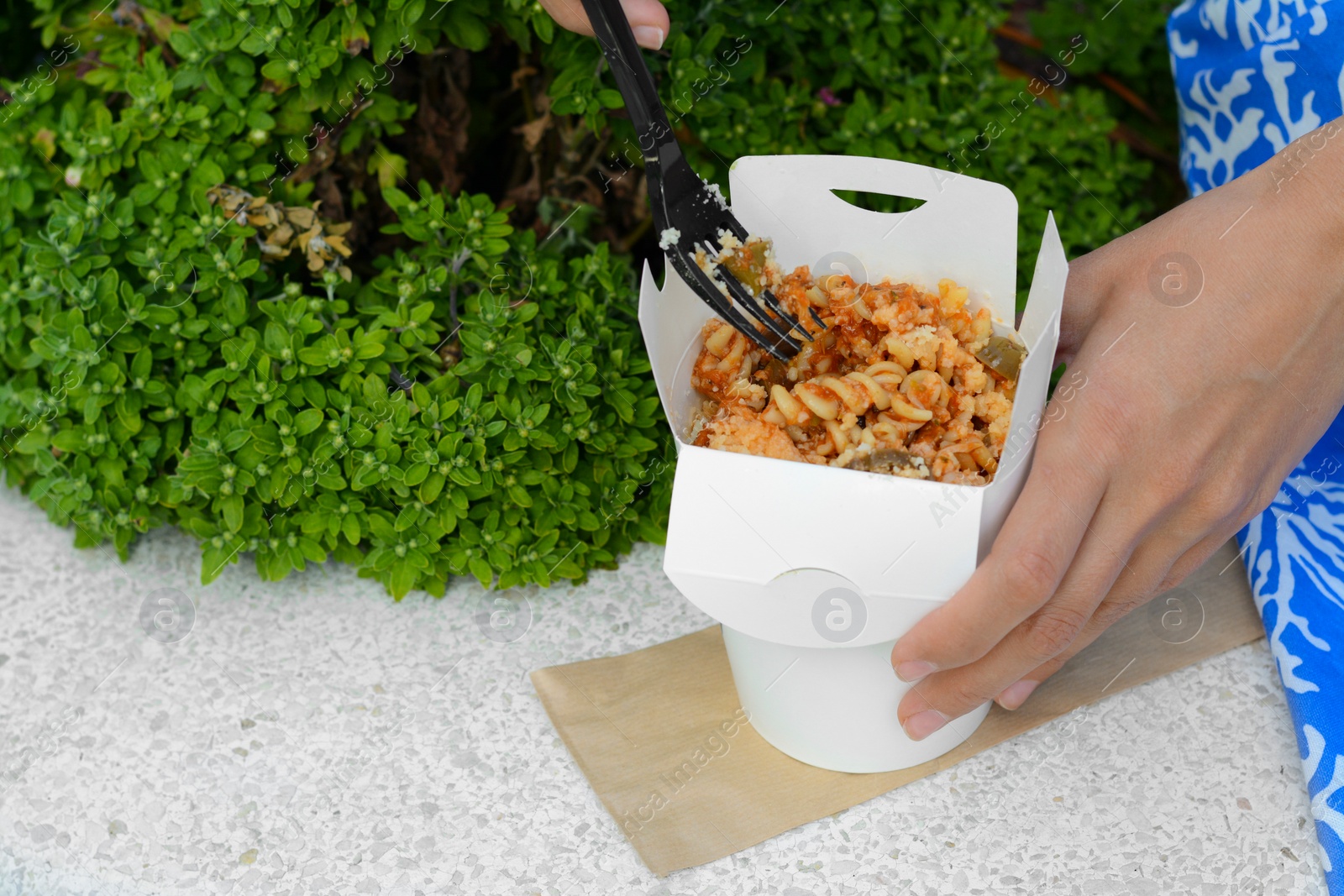 Photo of Woman eating takeaway noodles from paper box with fork on white surface outdoors, closeup and space for text. Street food