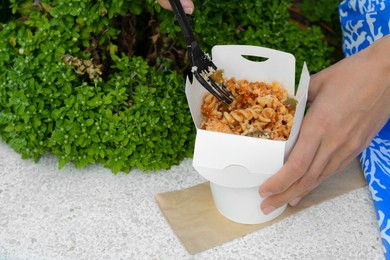 Woman eating takeaway noodles from paper box with fork on white surface outdoors, closeup and space for text. Street food